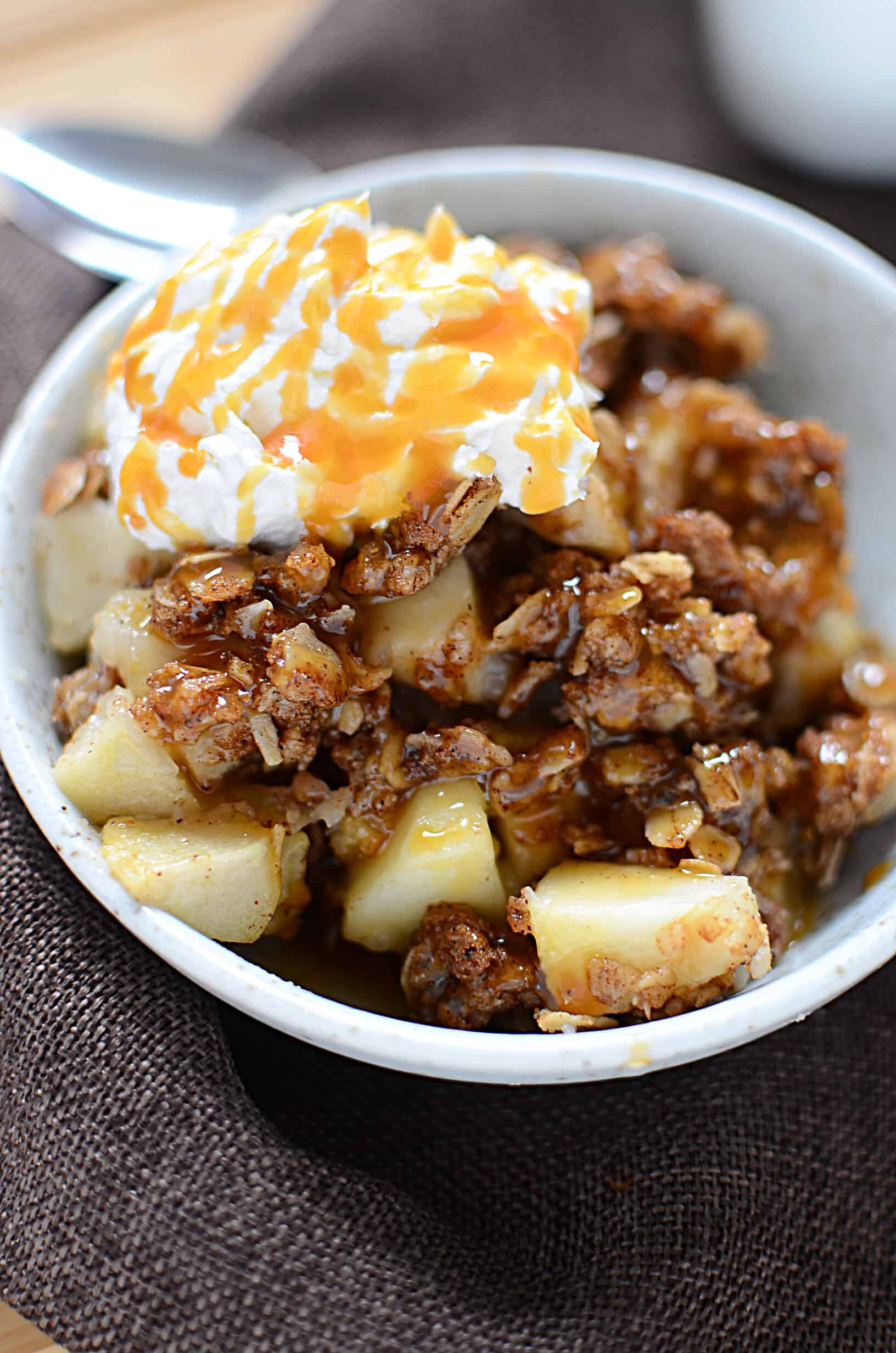 Spiced apple crisp with caramel drizzled on top in a white bowl with whipped topping.