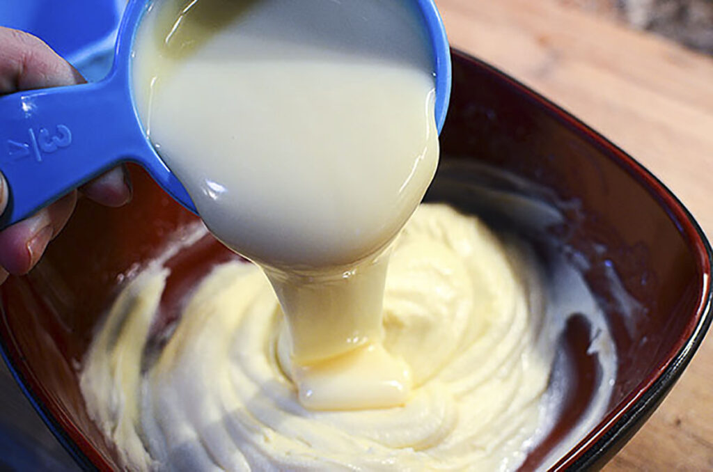 A blue measuring cup pours the sweetened condensed milk into the red mixing bowl.