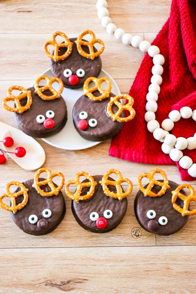 Six reindeer wagon wheel cookies on a wood table with a red napkin to the right and wooden bead garland on top.