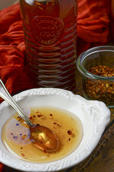 A white condiment bowl holds a bit of the honey with a spoon resting in it. Behind is the bottle of honey and hot chili peppers.