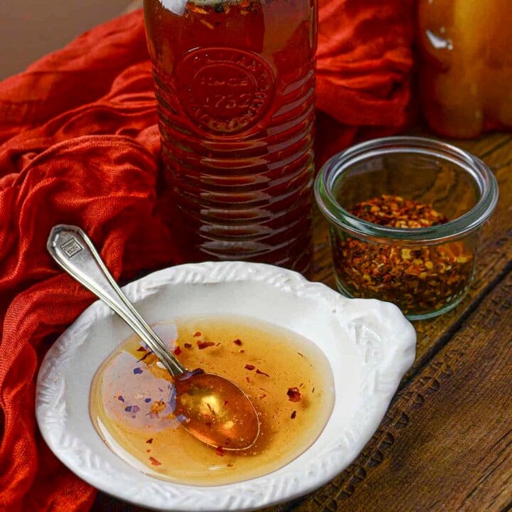 A white condiment bowl holds a bit of the honey with a spoon resting in it. Behind is the bottle of honey and hot chili peppers.