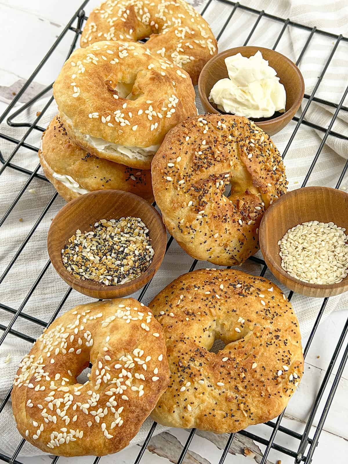 A variety of bagels on a cooling rack with bowls of seedy toppings in between them. 