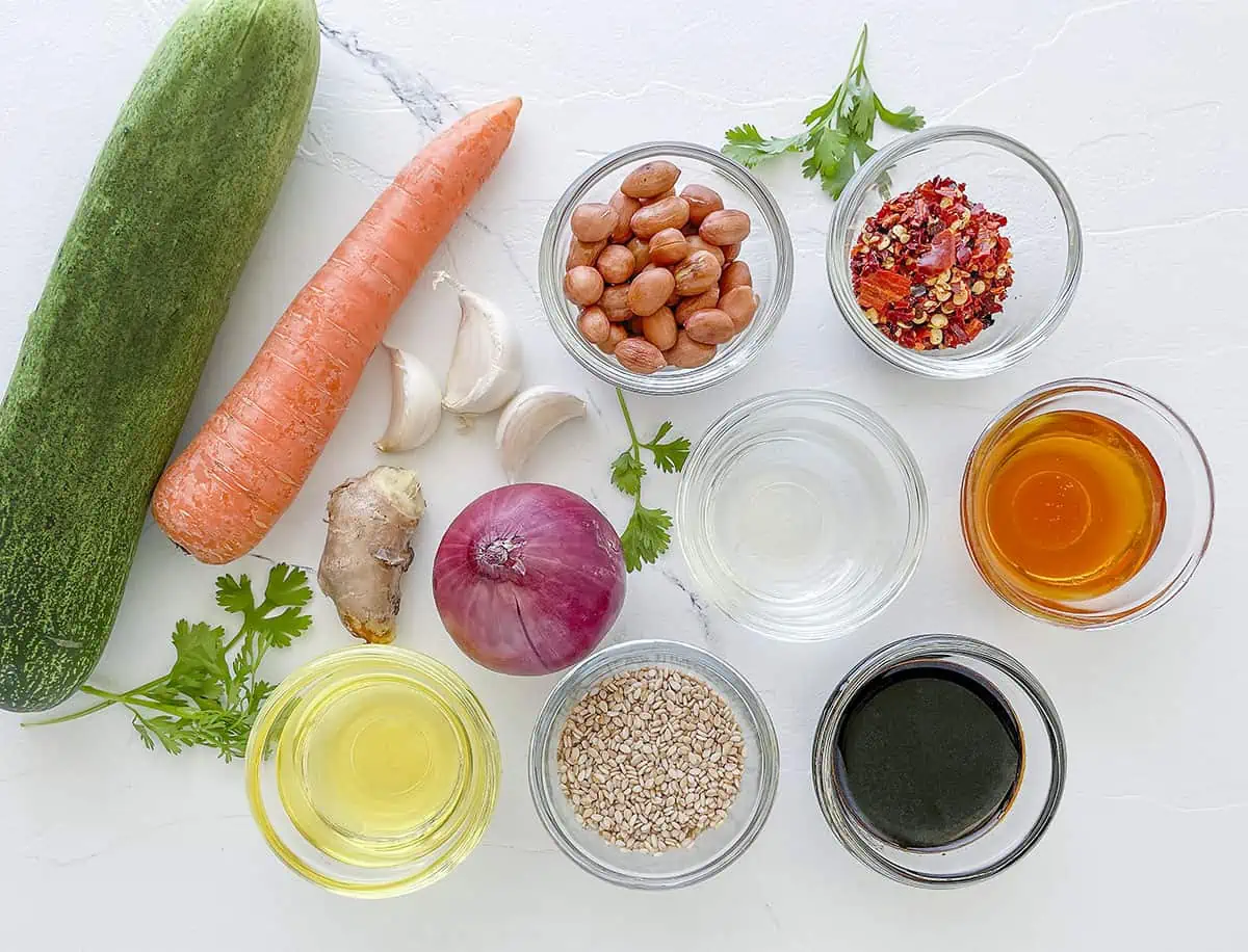 The ingredients for the salad are shown. From left to right, cucumber, carrot, garlic, peanuts, chilis, honey, vinegar, sesame seeds sesame oil, red onion ginger and cilantro. 