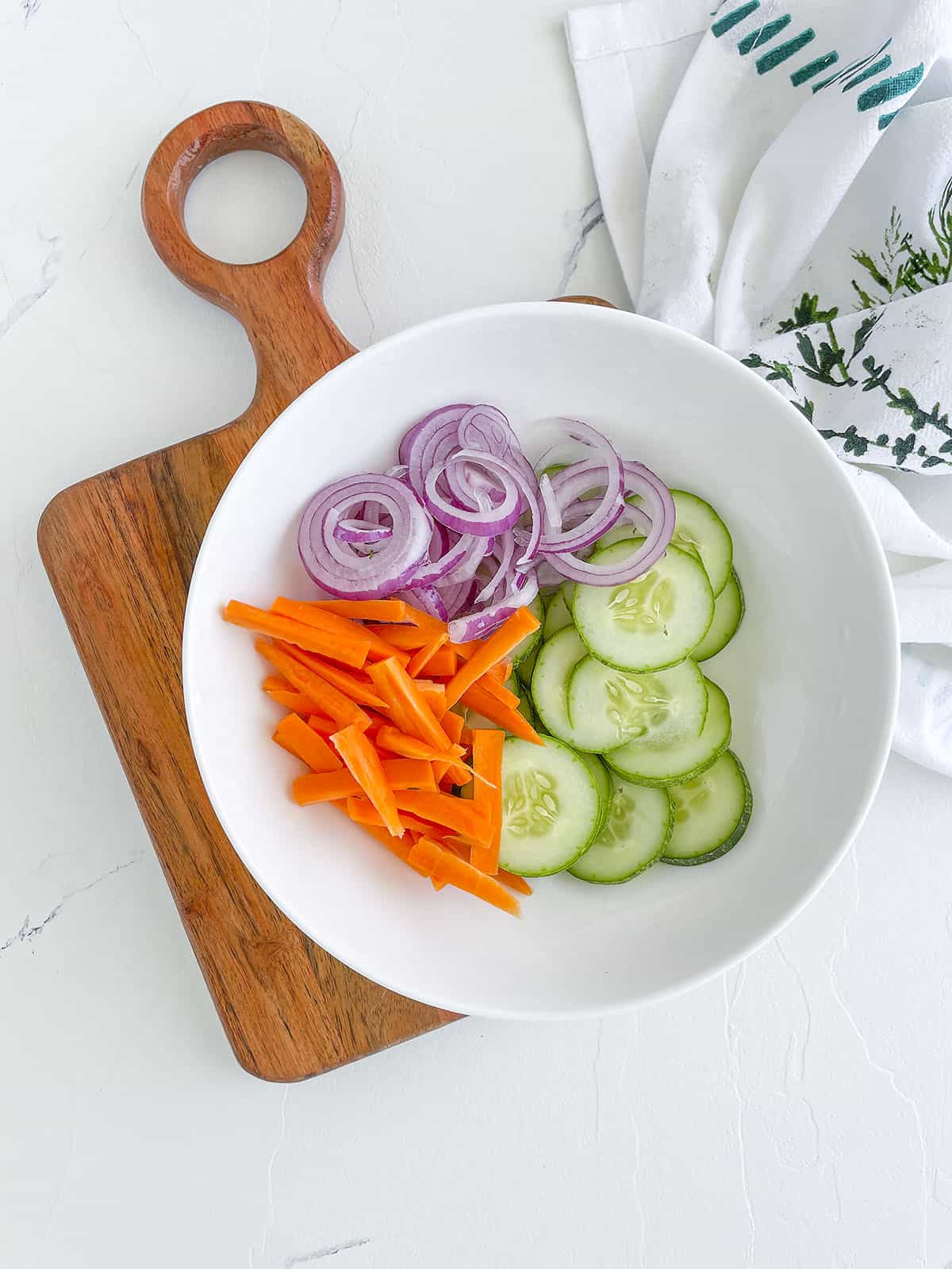 A wood cutting board with a white bowl holding cut carrots, red onions and sliced cucumbers. 
