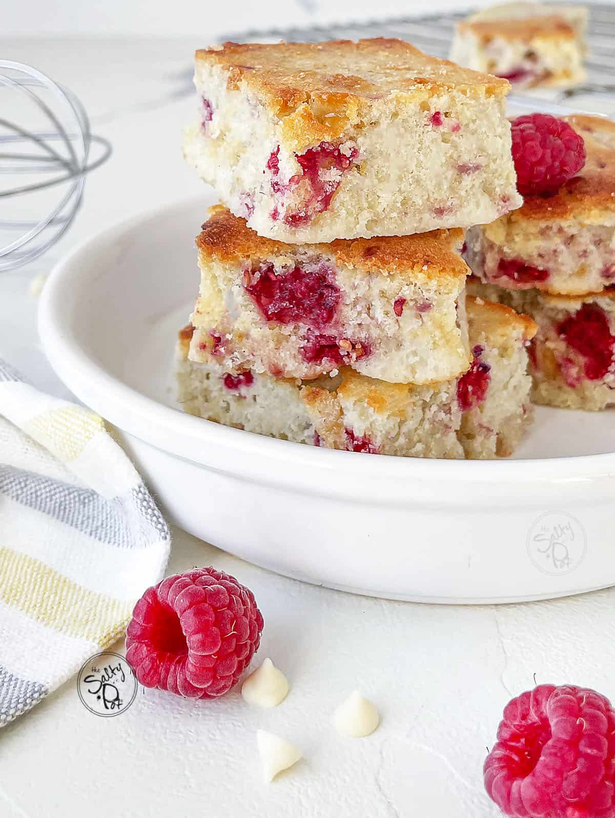 Raspberry White Chocolate Blondies in a white ceramic bowl with a raspberry up front of it.