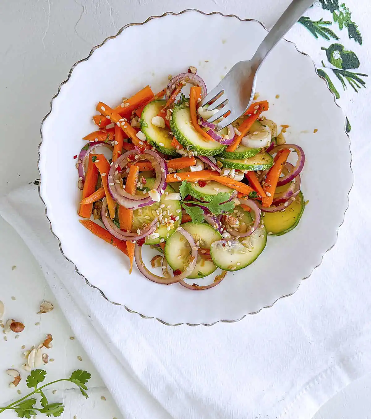 A fork is peircing  a slice of cucumber and carrot from the salad that's in a white bowl. 