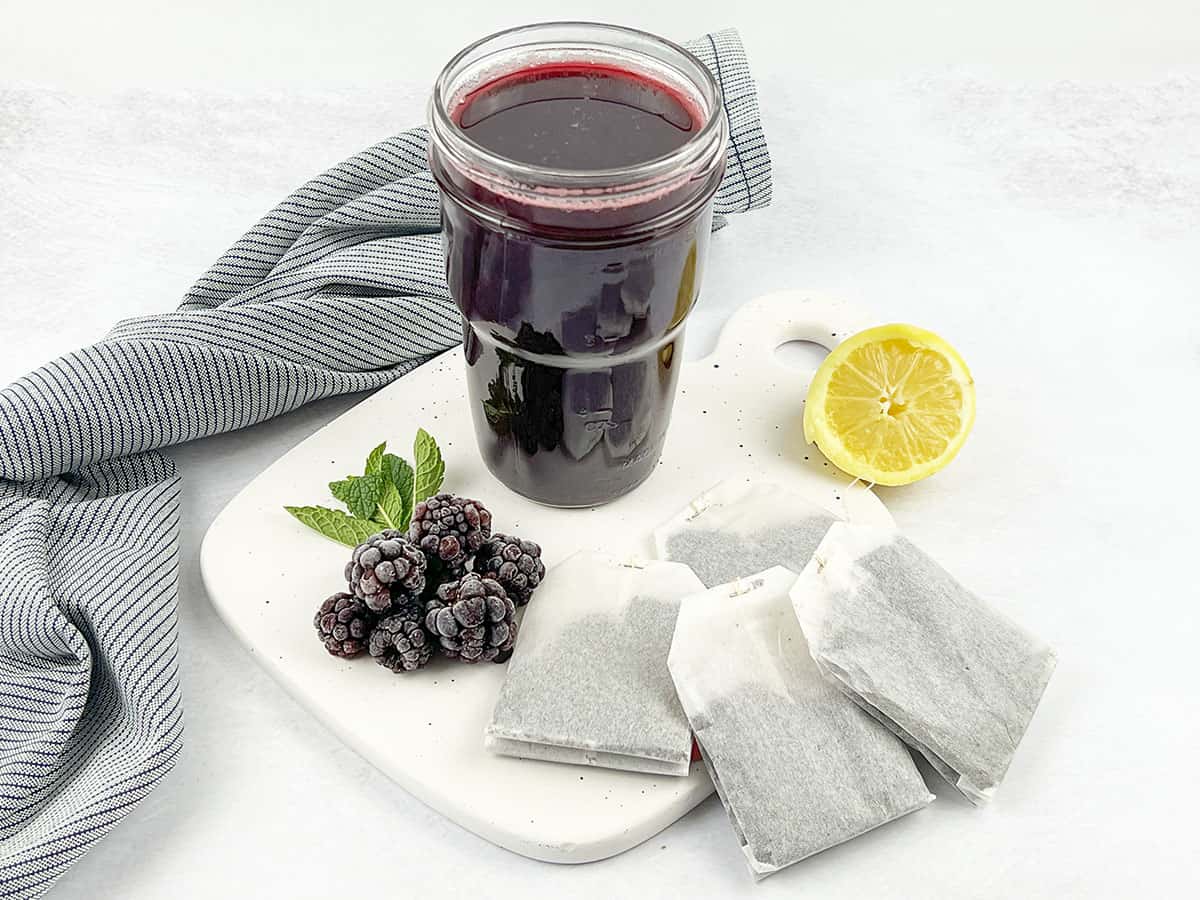 Black simple syrup in a large glass with lemon wedges and blackberries beside it. Tea bags lay on the ceramic cutting board.