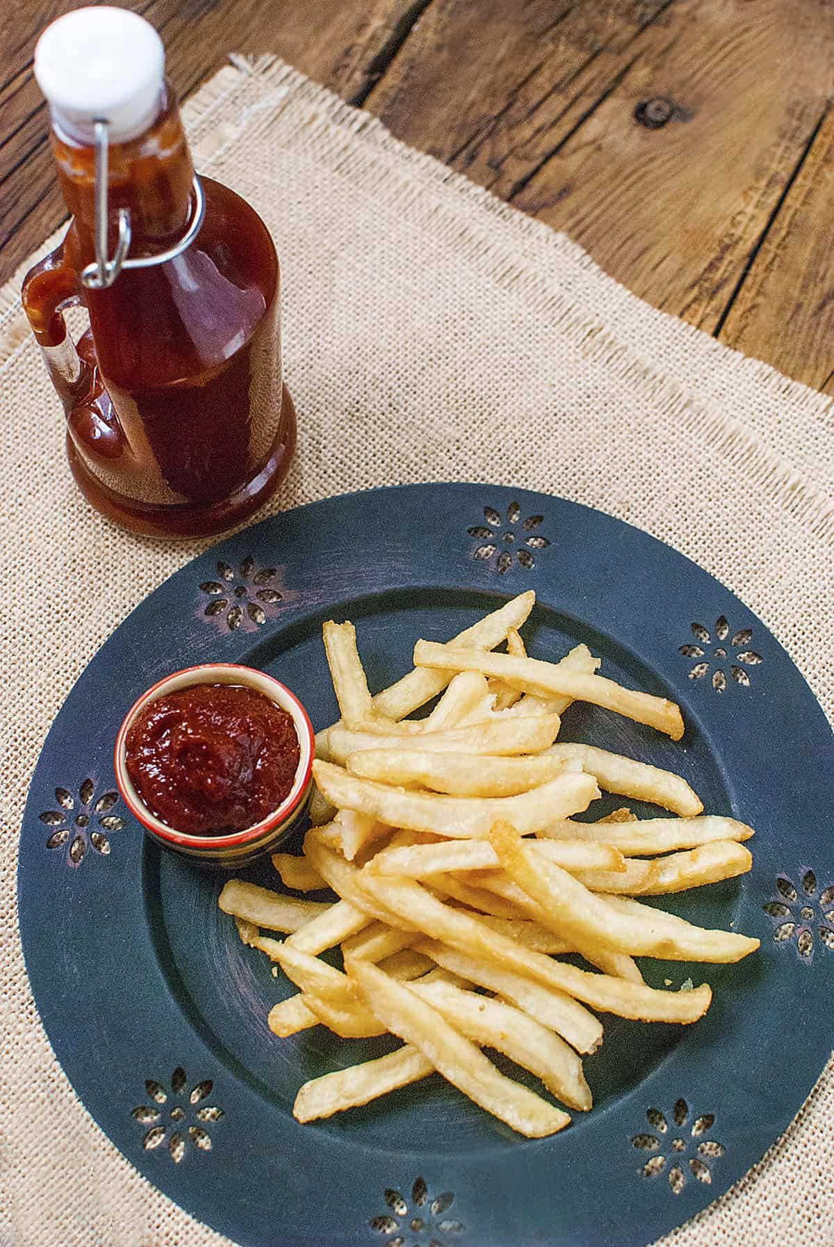 A bottle of slow cooker ketchup next to a plate of french fries. There is a small bowl of ketchup on the plate.