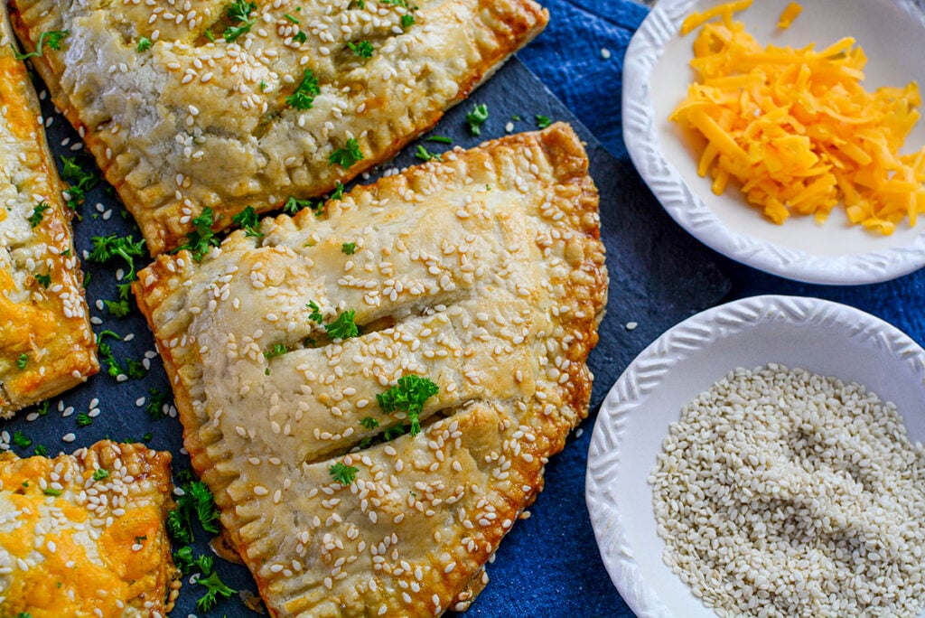 One chicken and asparagus hand pie with no cheddar topping. There are two small bowls one holding shredded cheddar and another, sesame seeds.