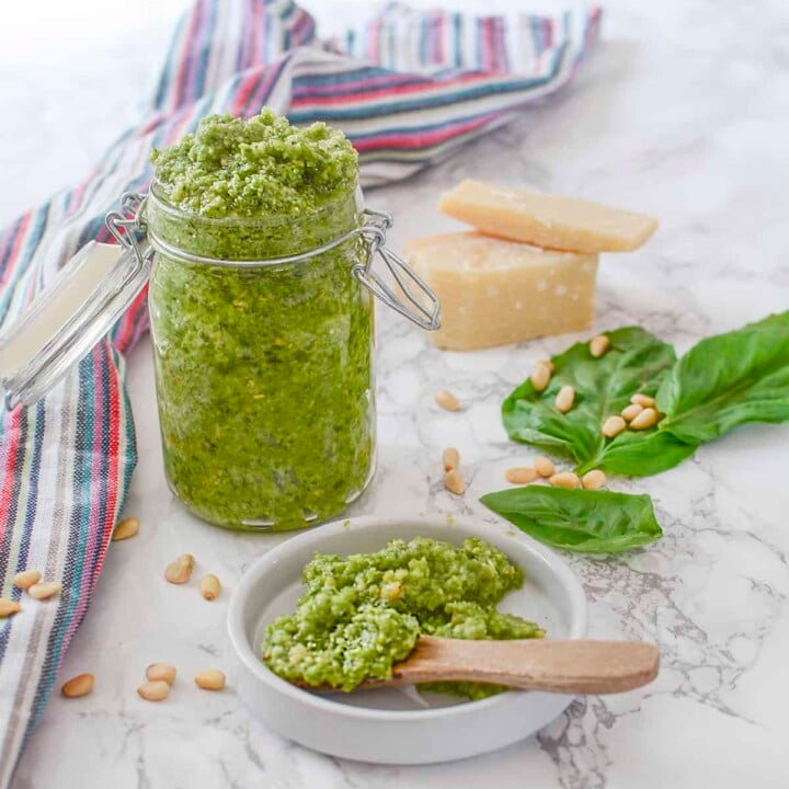 Garlic scape pesto in a tall jar with some in a white plate in the foreground.