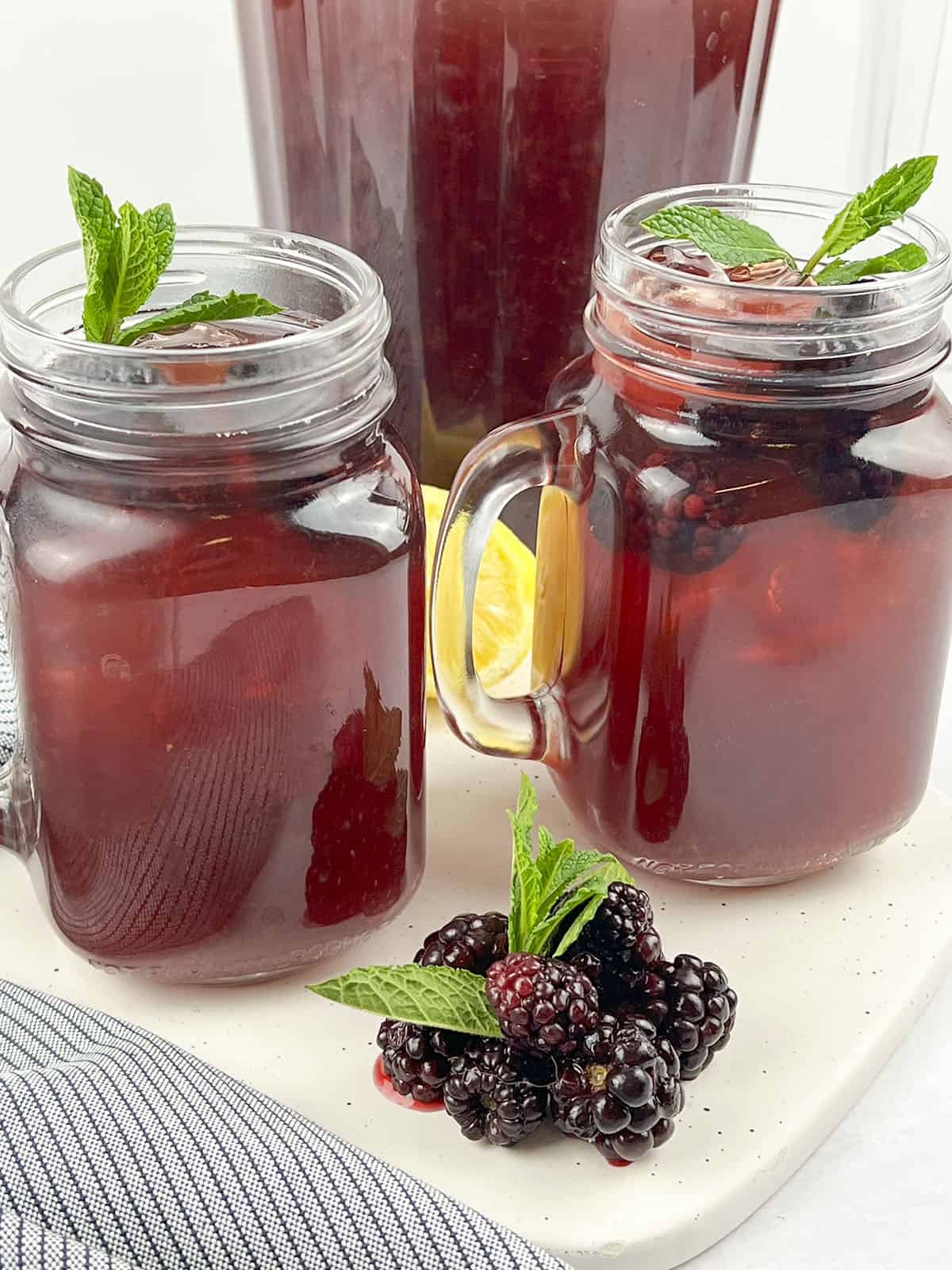 Two glasses of iced tea with a pitcher in behind them. In front there is a small pile of blackberries with mint.