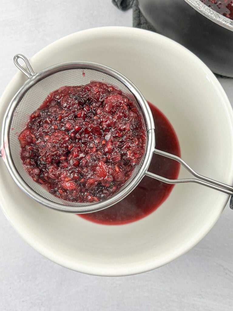 The blackberries being strained through a sieve over a large white bowl.