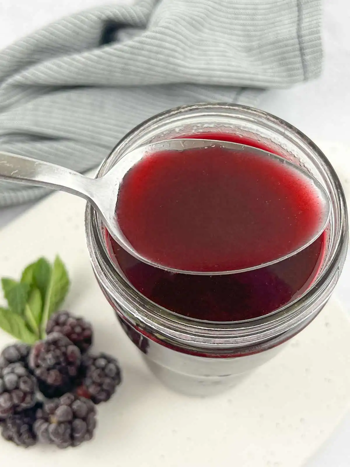 A spoonfull of blackberry syrup over the glass jar with blackberries and a grey napkin in the background.