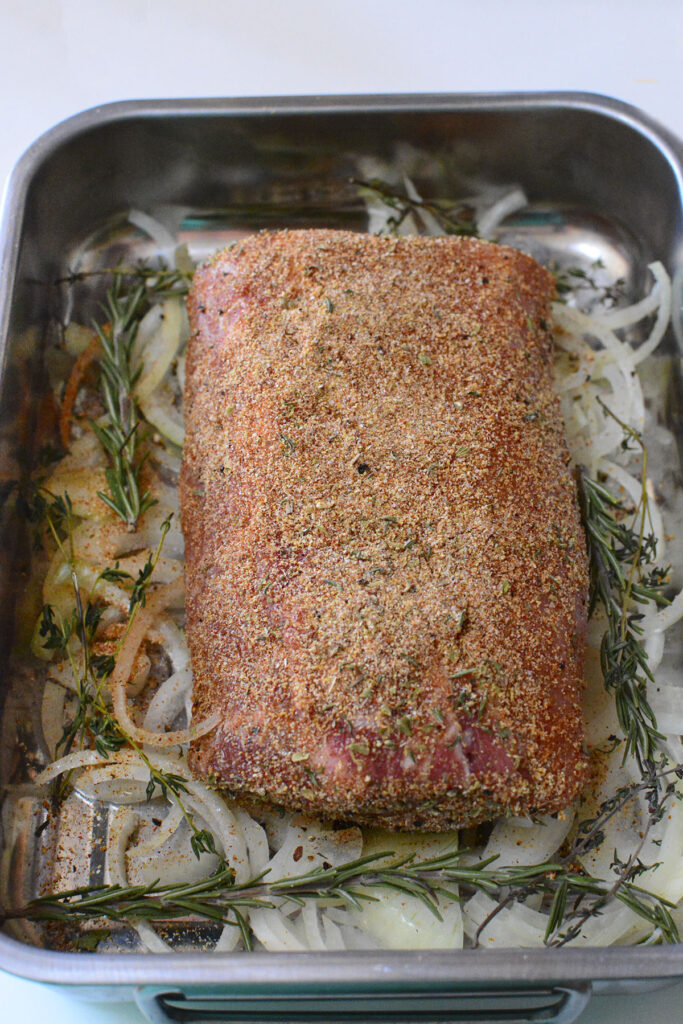 The seasoned pork loin resting on top of the bed of onions, with fresh rosemary and thyme on the sides.