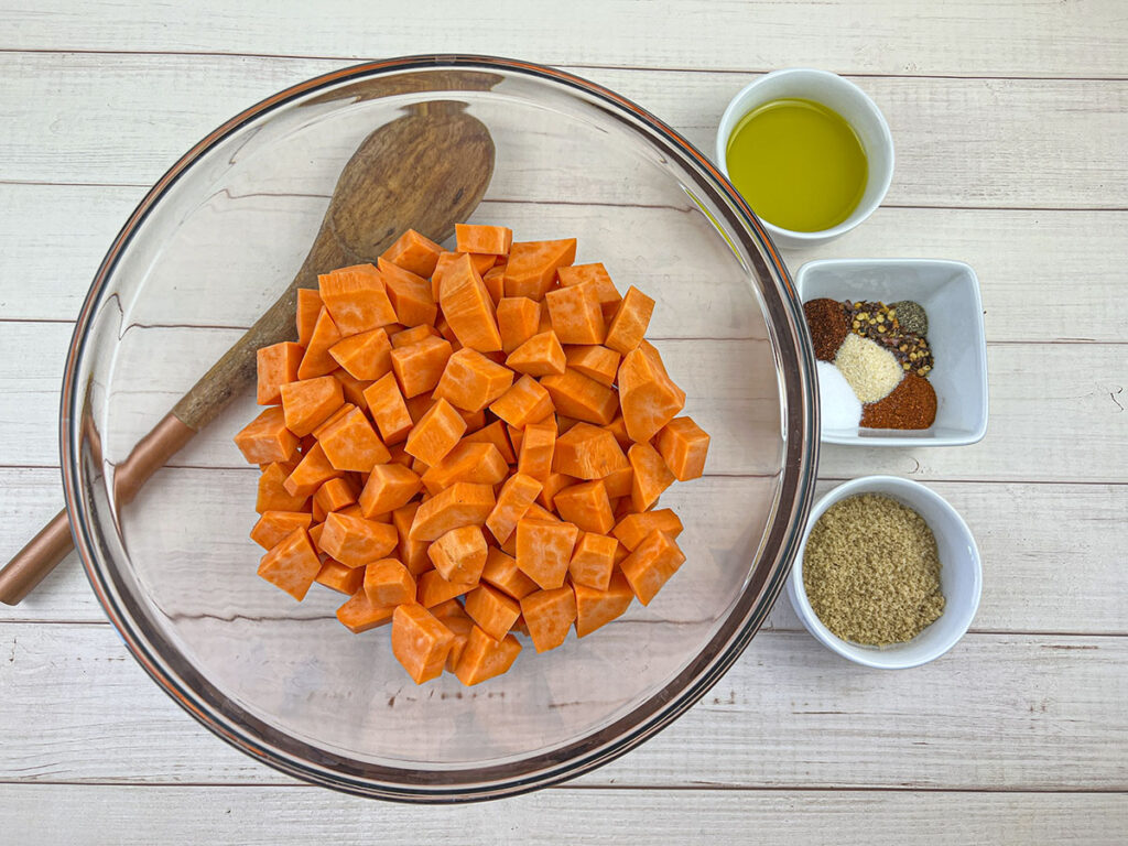 Sweet potatoes in a glass bowl with seasonings and oil on the right, ready to go in the bowl.
