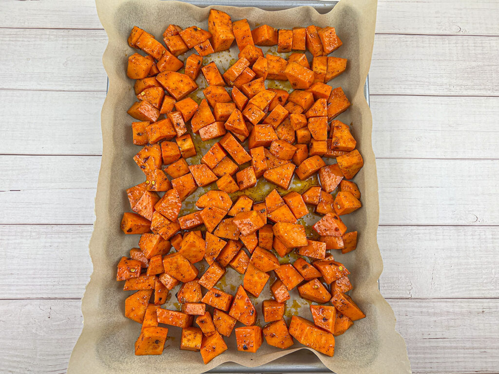 Seasoned raw sweet potatoes on a parchment lined baking sheet ready to go in the oven.