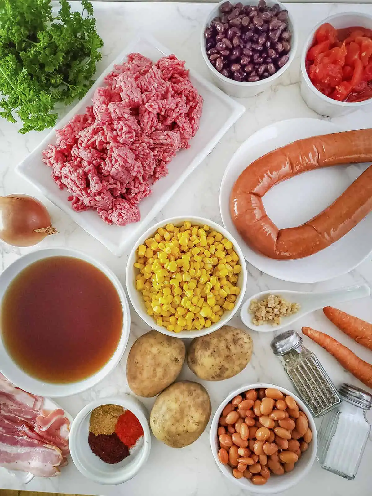 Ingredients for the cowboy soup. Things like ground beef, beans, corn, potatoes and sausage, along with broth all sit on a white marble table. 