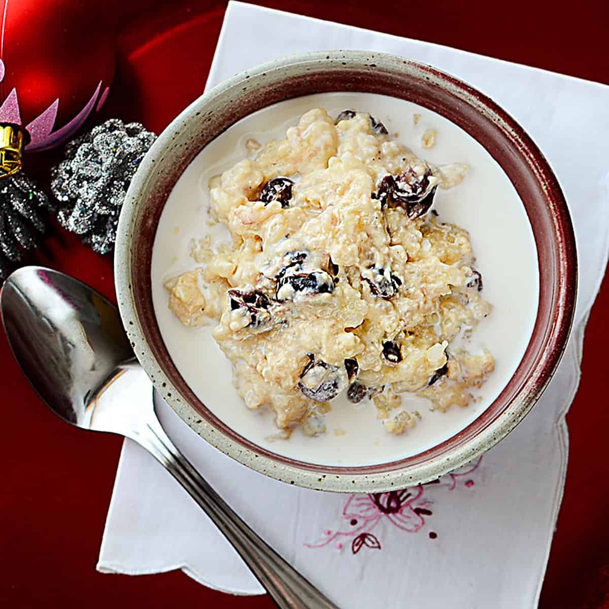 nstant Pot Eggnog rice pudding with cranberries in a red and cream pottery bowl with a cream colored napkin underneath. Square image.