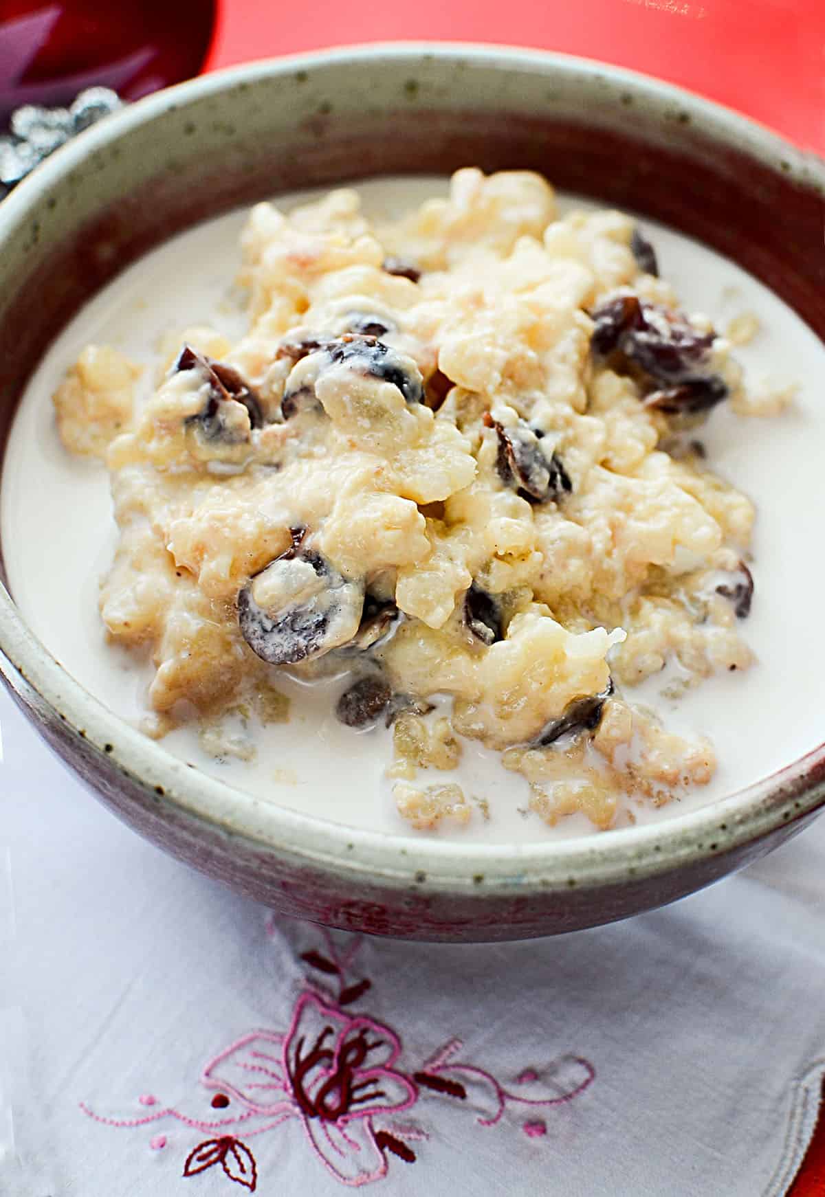 A close up photo of eggnog rice pudding with cranberries, served with cream in a pottery bowl. 