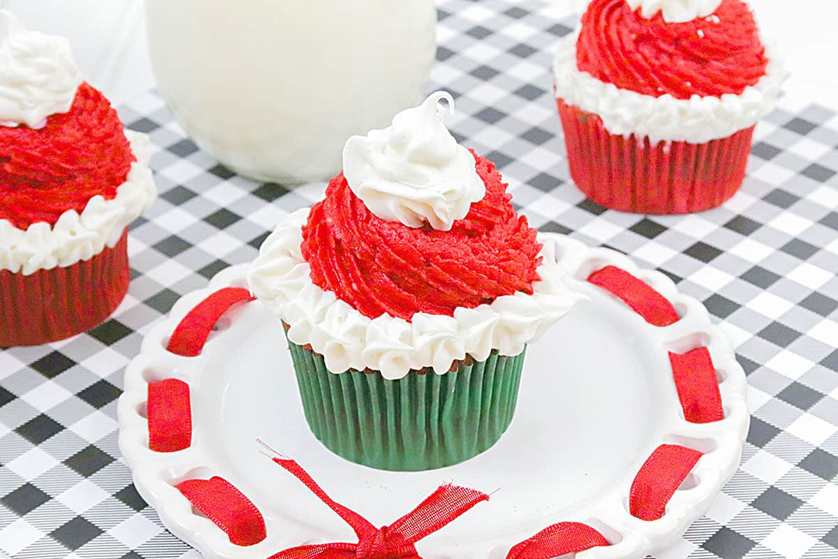 Three santa's hat cupcakes on a black and white checkered table cloth. A container of milk is in the background.