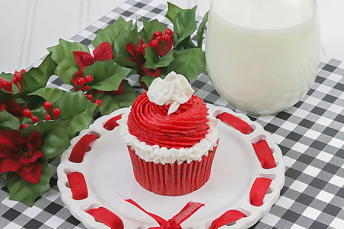 One Christmas Cupcake on a white plate with a big glass of milk on the right, and a sprig of holly on the top left.