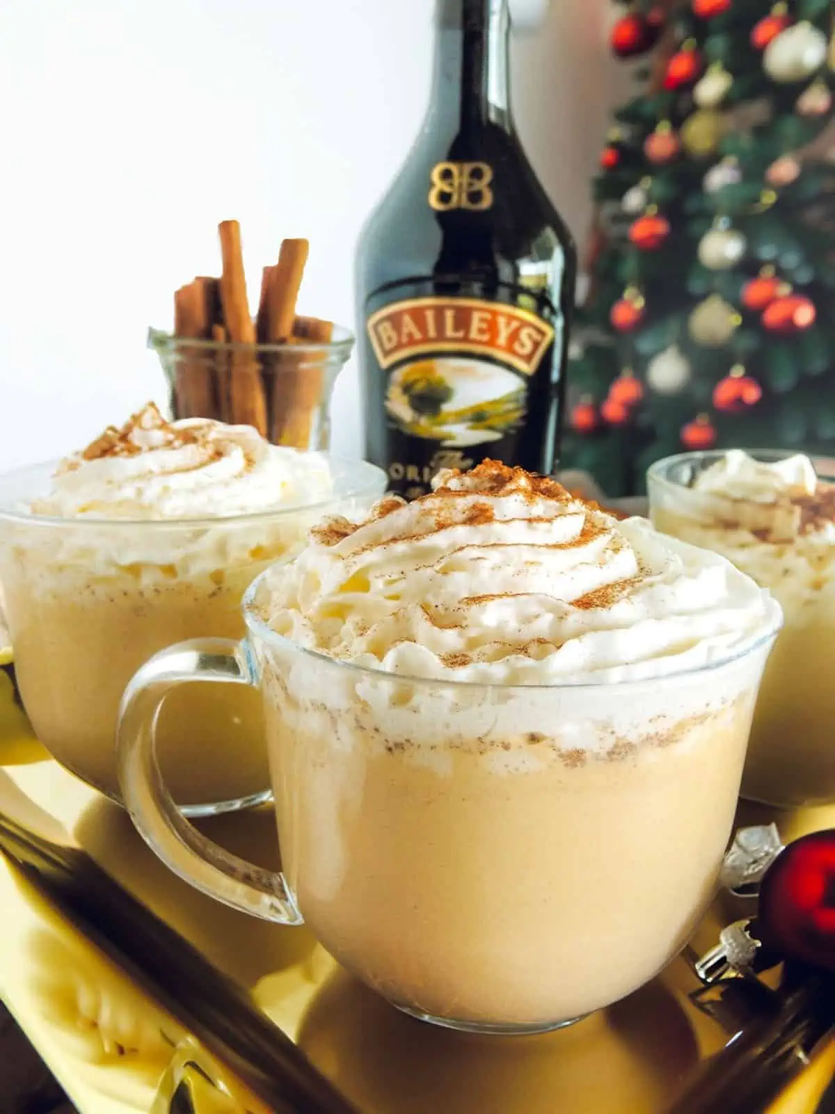 Baileys Eggnog made in three glasses on a gold tray. There is a christmas tree on the right in the background, with the bottle of baileys in the center and some cinnamon sticks on the left.