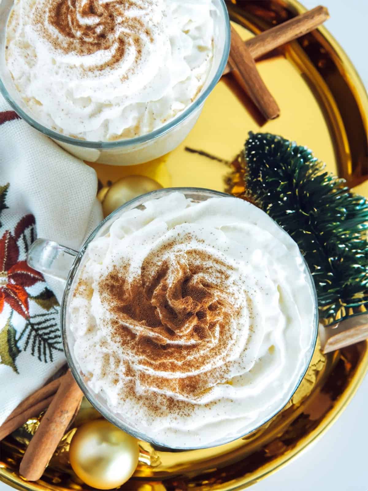 Two eggnog with baileys drinks with whipped cream and cinnamon on top. There is a tiny christmas tree on the right and a festive napkin on the left.