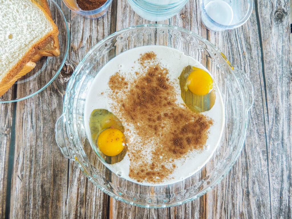 Eggs, milk, baileys and cinnamon in a dish. Slices of bread are up in the upper left corner.