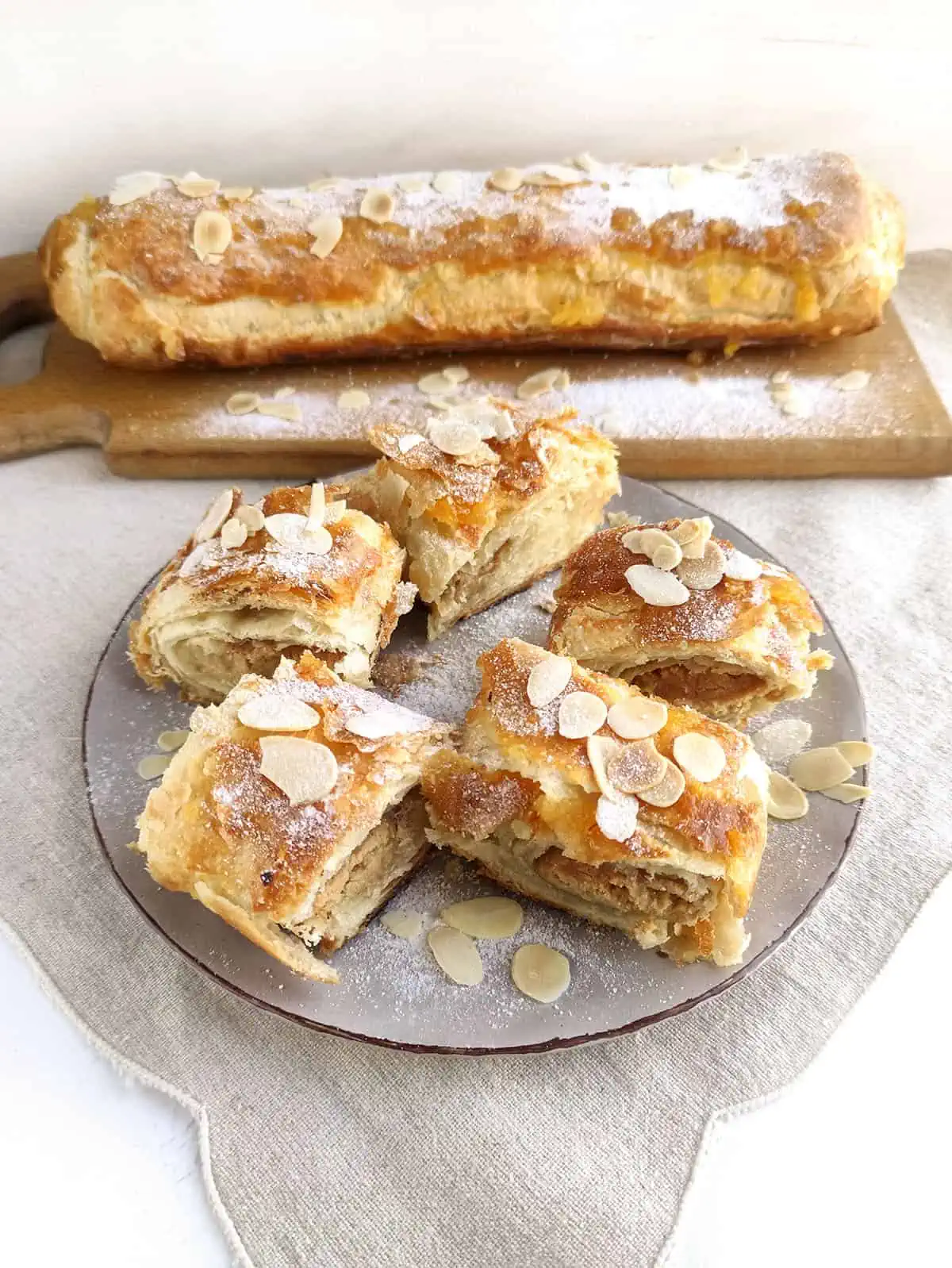 5 pieces of Almond Banket on a plate with flaked almonds on top with icing sugar. There is a uncut log of banket in the background.