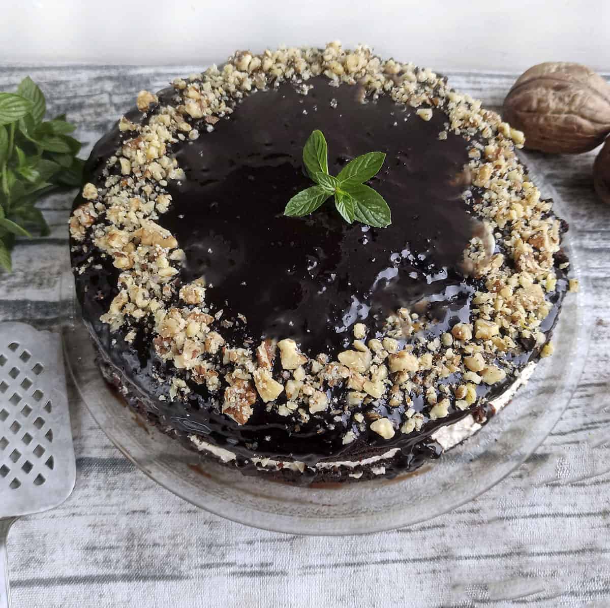 Chocolate cake with walnuts on a clear plate. A cake server is on the left side of the photo.