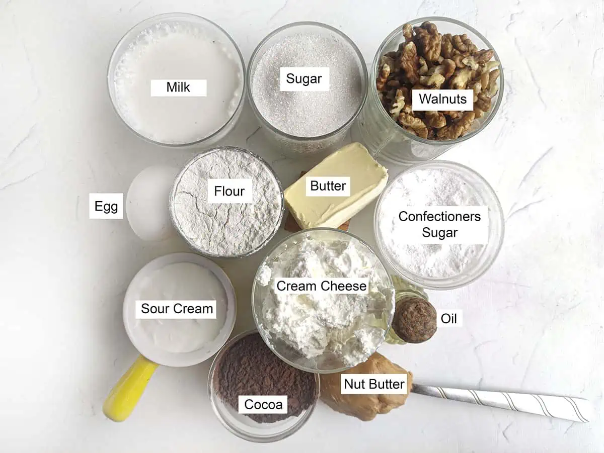 Some of the ingredients needed to make this chocolate walnut cake. Starting from the top left, we have Milk, sugar, walnuts, egg, flour, butter, confectioners sugar, sour cream, cream cheese, oil, cocoa, and nut butter.