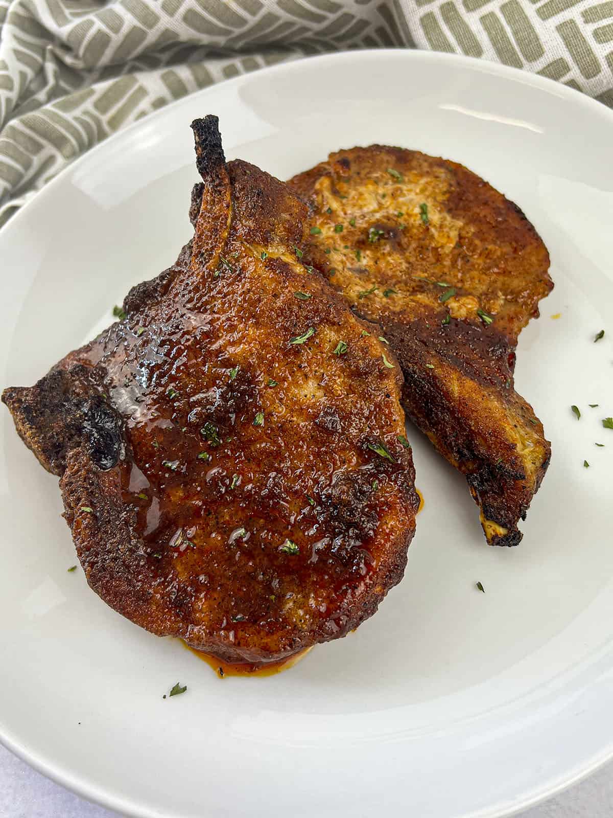 Two pork chops that are seasoned and air fried sitting on a white plate with a napkin in the background.