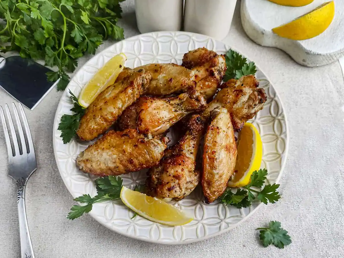 8 chicken wings on a white plate with lemon slices. There is a fork to the left of the plate with green parsley at the top left.