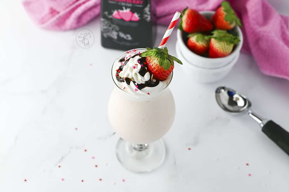 Garnished with whipped cream, chocolate drizzle and a fresh strawberry, the cocktail sits with a white bowl of strawberries next to it. 