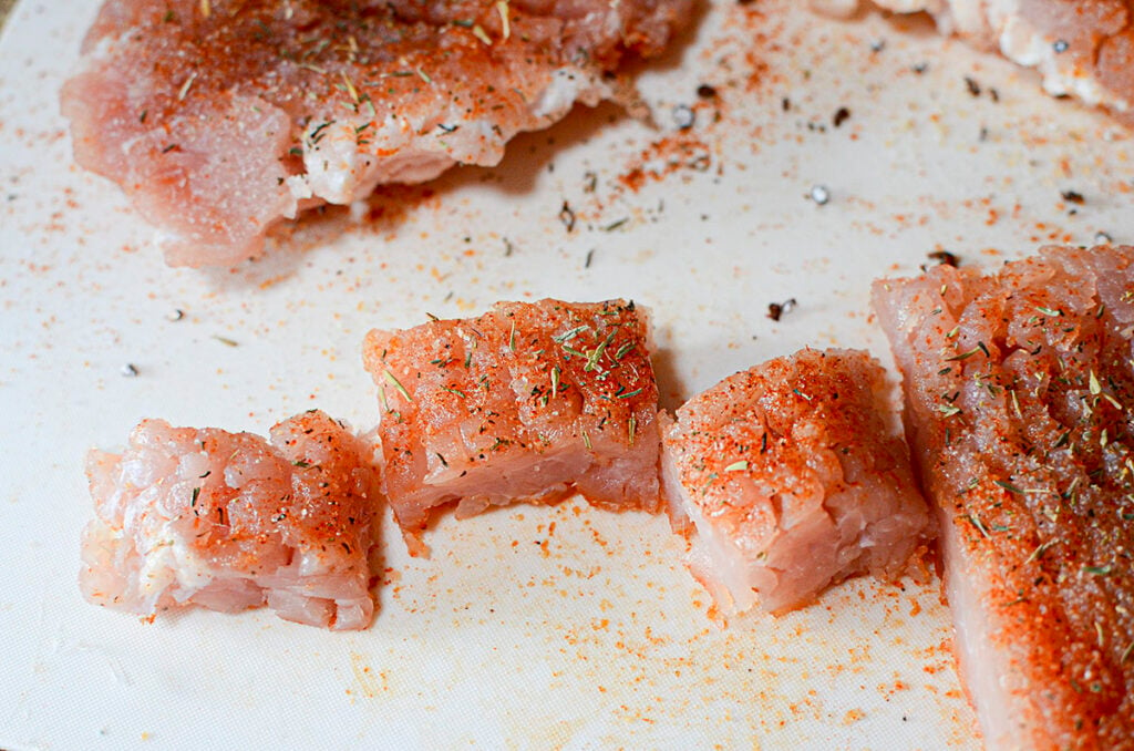 Raw pork that's been seasoned, cut into cubes on a white cutting board.
