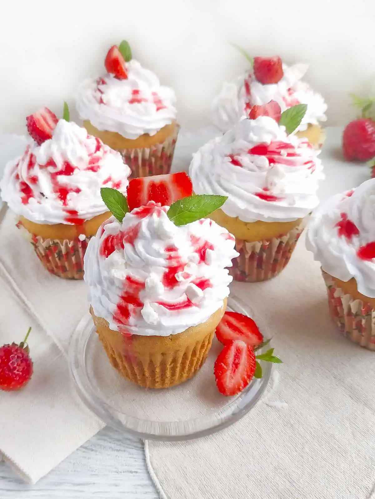 6 cupcakes with whipped topping and strawberry puree drizzled on top on a linen table cloth.