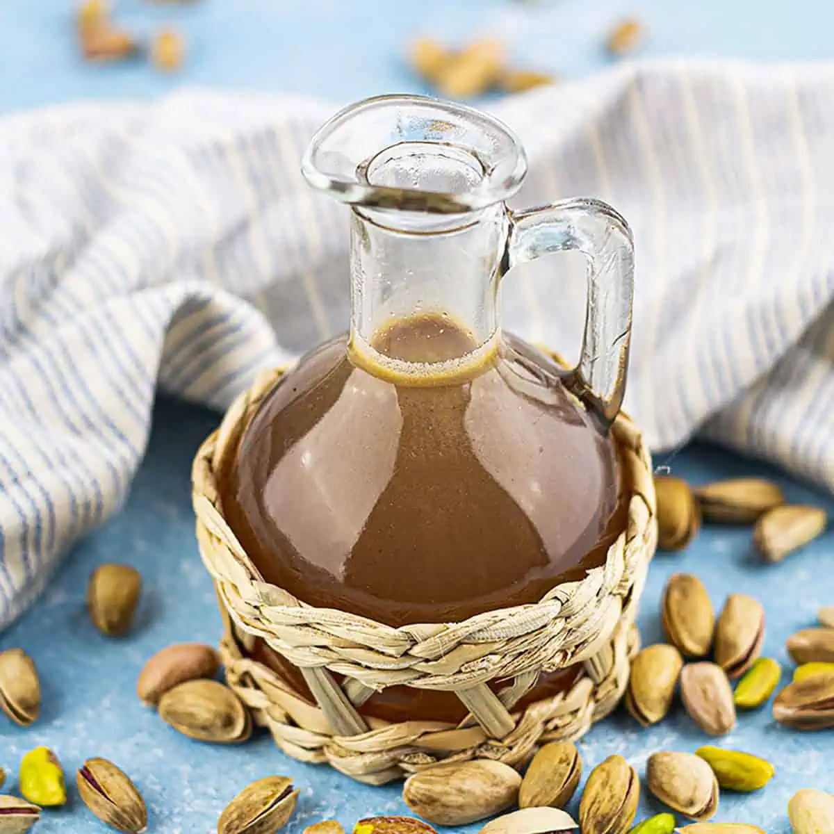 The pistachio syrup in a glass carafe with a bamboo decoration. Pistachios sit on the table around it. Square image.