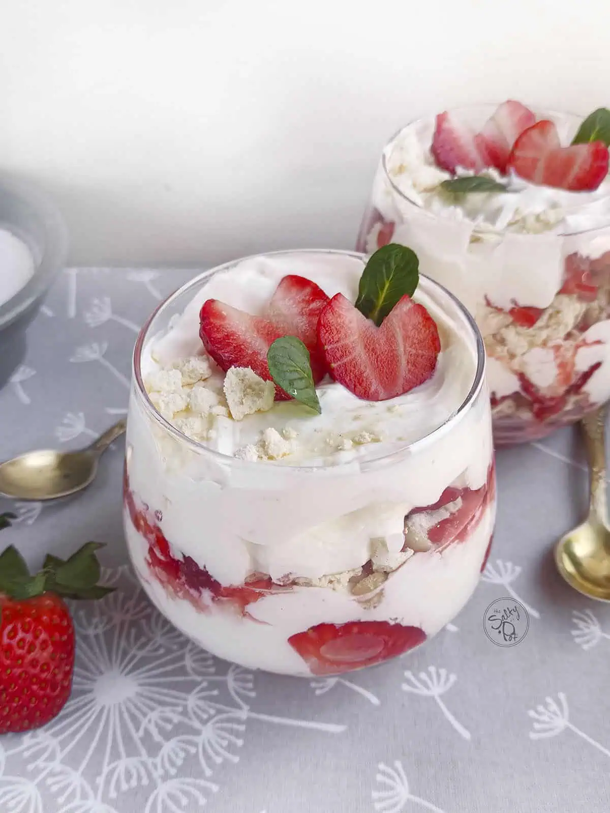 2 Strawberry Eton Mess parfaits in dessert glasses with two spoons on a grey tablecloth.