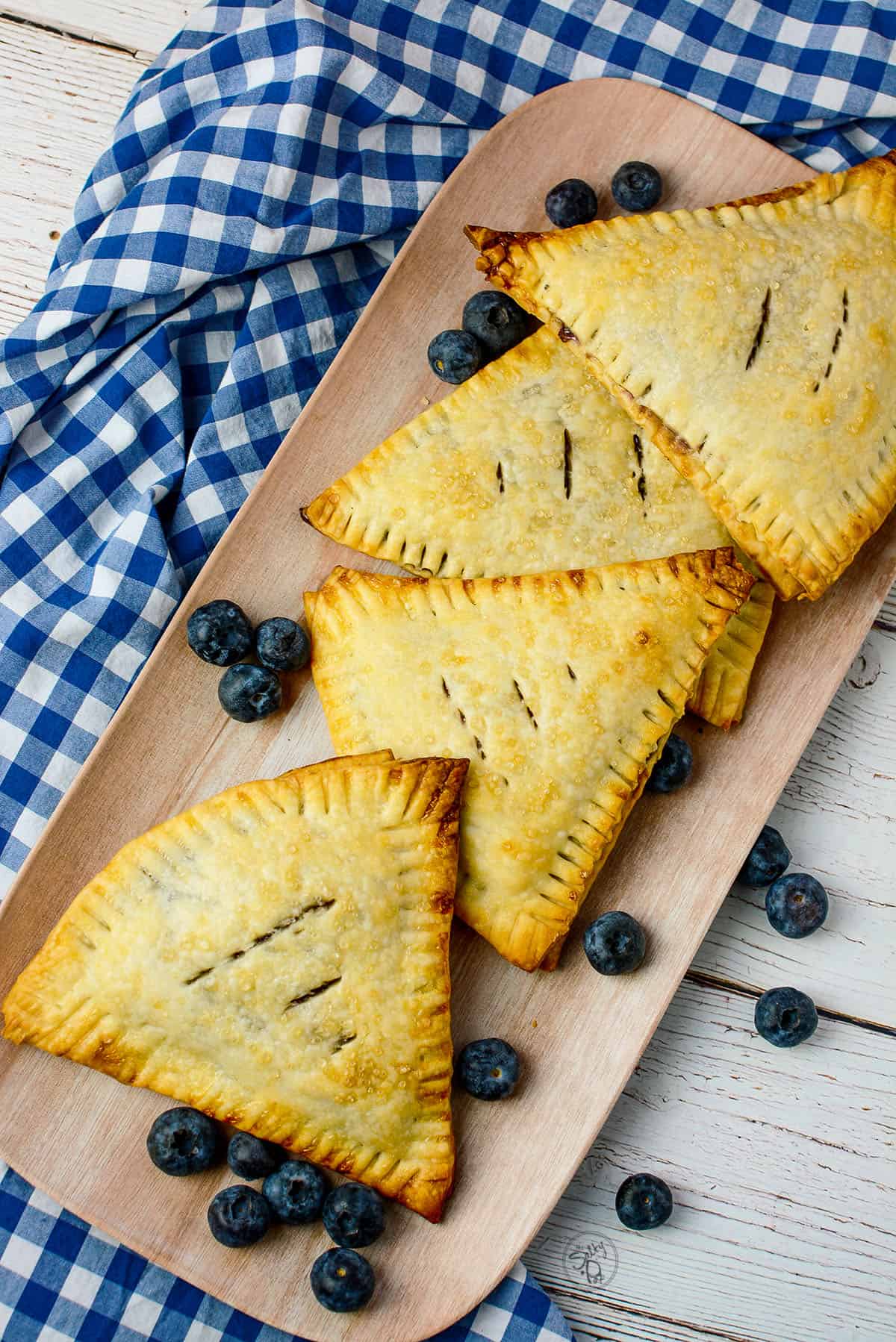 Four blueberry hand pies on a tray with a blue and white checkered cloth next to the plate on the left.