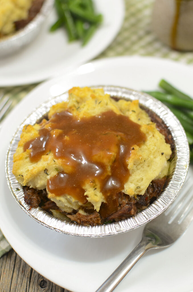 Individual beef pot pie with biscuit topping with gravy drizzled over top. Green beans are on the right side.