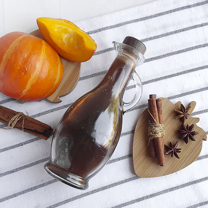 Pumpkin spice coffee syrup in a jar laying on it's side. Square image. Pumpkin wedges on the upper left.