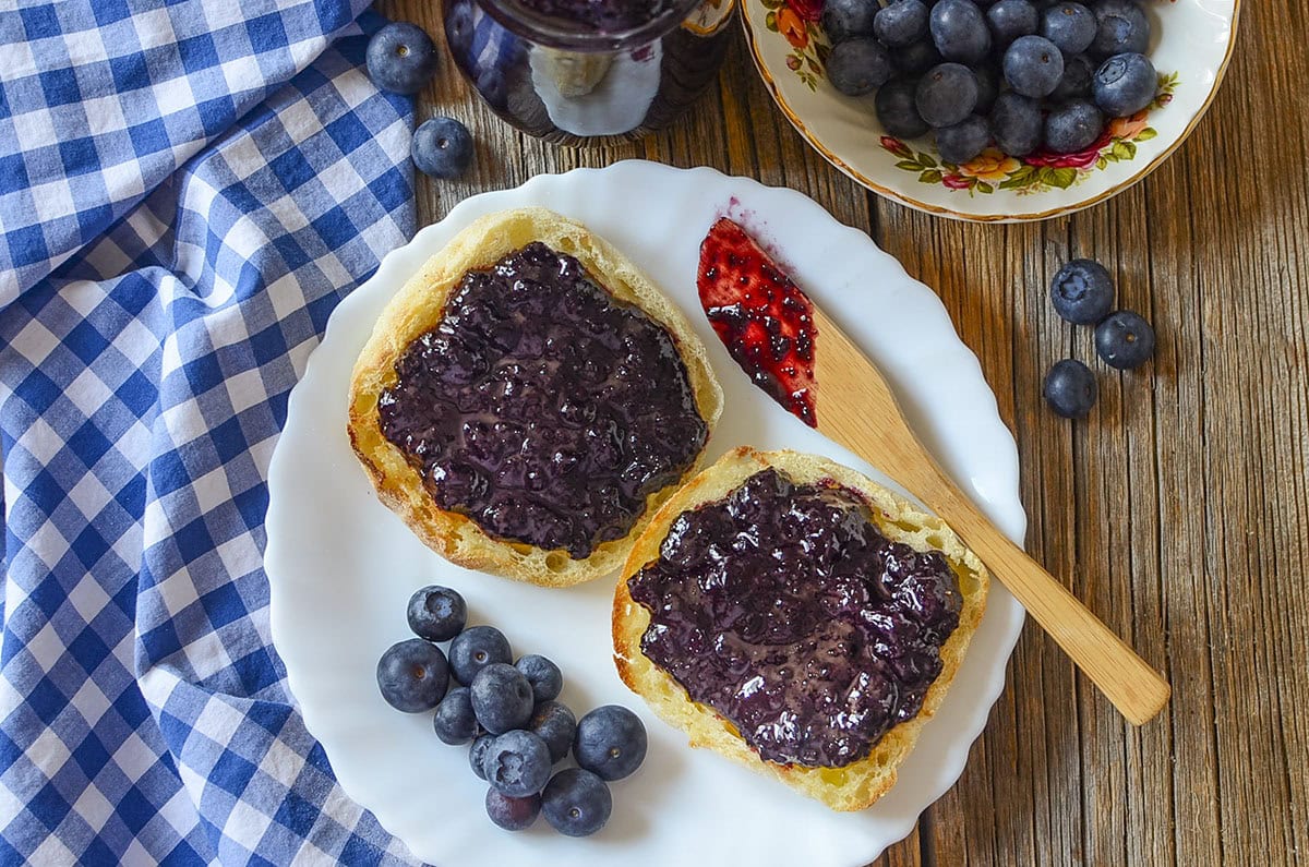 Blueberry butter spread on english muffins on a white plate with a wooden knife on the right.