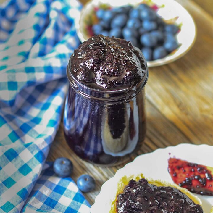 Blueberry butter in a glass jar with fresh blueberries in the background on the top right.