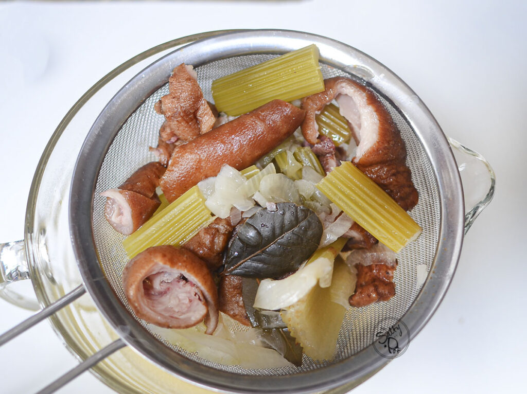 A seive filled with cooked soup ingredients for stock. Smoked pork hock, bay leaf, celery, onions, etc.