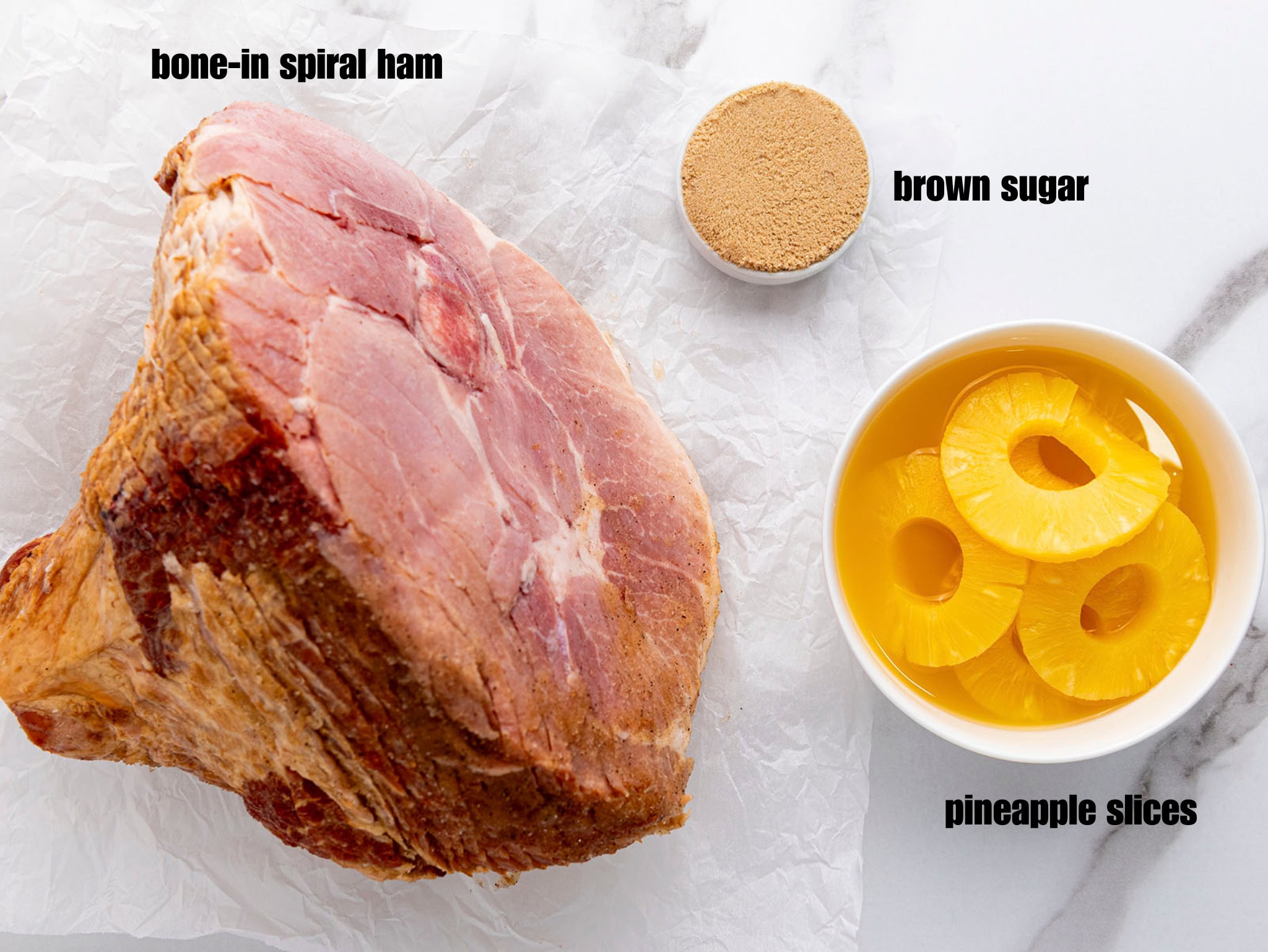An overhead view of ingredients set on a crinkled parchment paper and marble counter. A large, bone-in ham sits on the paper. Below it are two small, white bowls: one containing yellow pineapple slices in juice, and the other containing brown sugar.