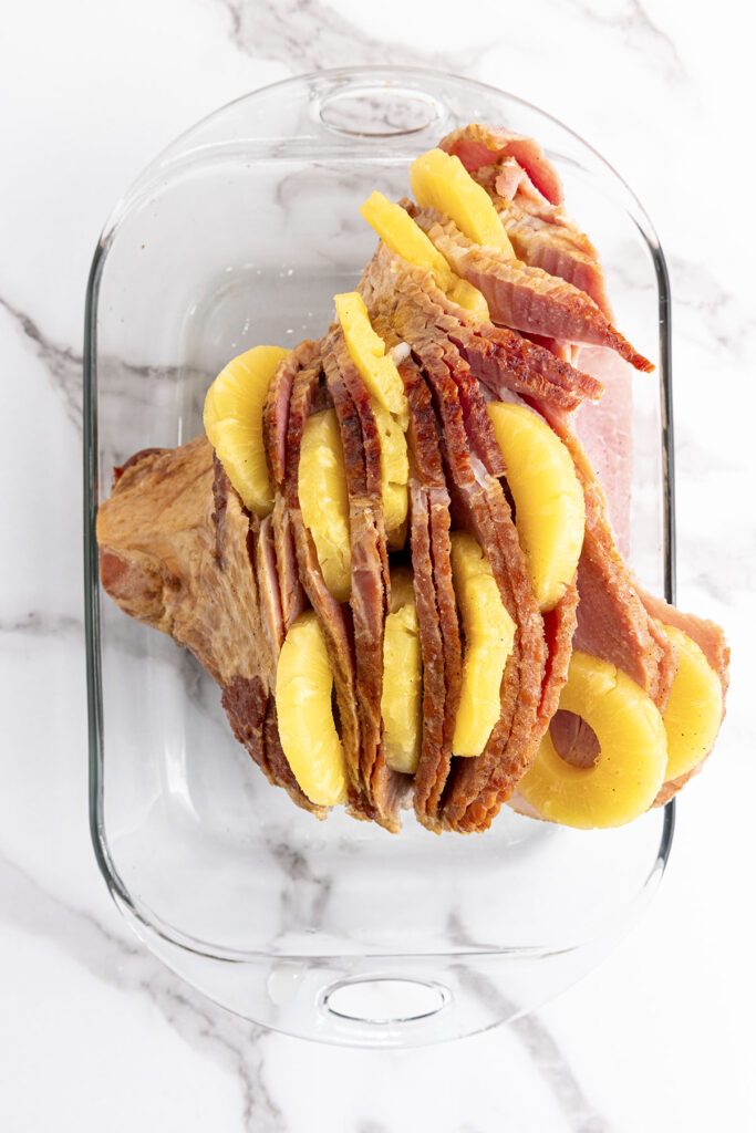 A top-down photograph looking into a clear glass, oval casserole dish on a white marble surface. A whole, pre-sliced spiral ham rests in the dish. Bright yellow pineapple slices are actively being tucked and shoved between the natural layers of the ham meat.