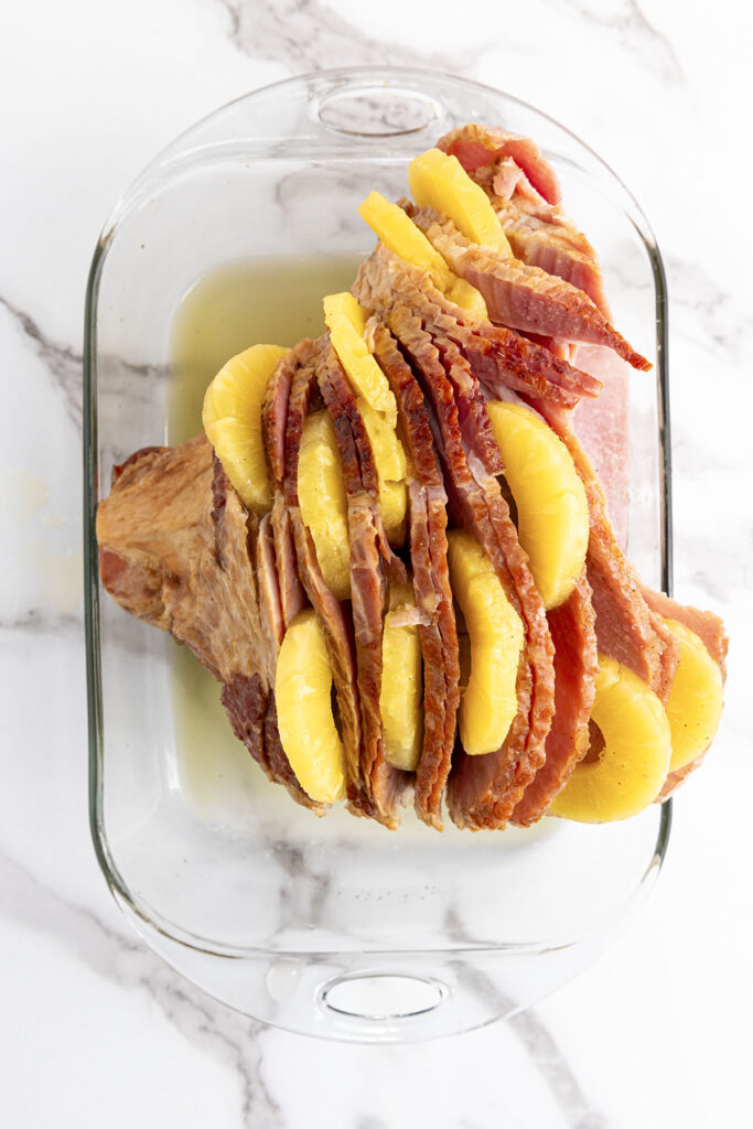 A top-down photograph looking into a clear glass, oval casserole dish on a white marble surface. A whole, pre-sliced spiral ham rests in the dish. Bright yellow pineapple slices are actively being tucked and shoved between the natural layers of the ham meat with pineapple juices at the bottom