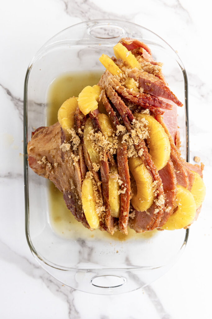 An overhead view of the ham in the oval glass casserole dish. The pineapple slices are already tucked between the layers. The whole ham is covered in a substantial layer of crumbled brown sugar and pineapple juice. The bottom of the dish is wet with the accumulated liquid.