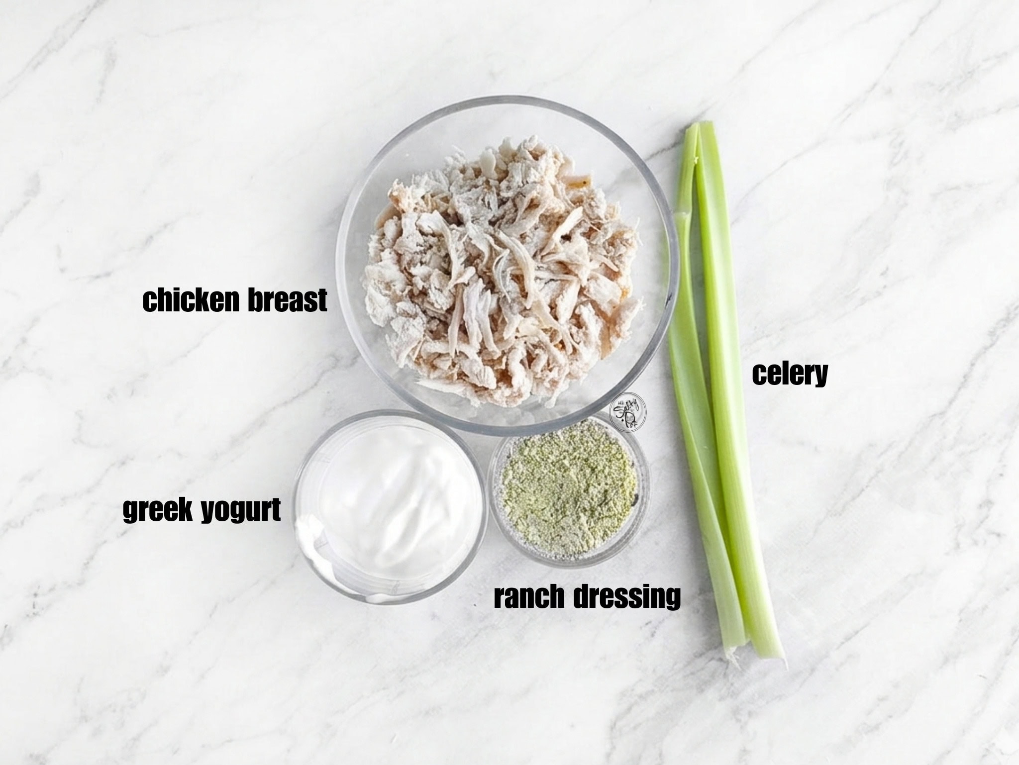 Overhead shot of the prep station featuring a glass bowl of shredded chicken, a bowl of thick Greek yogurt, a small dish of green herb ranch seasoning, and two fresh celery stalks on a white marble surface.