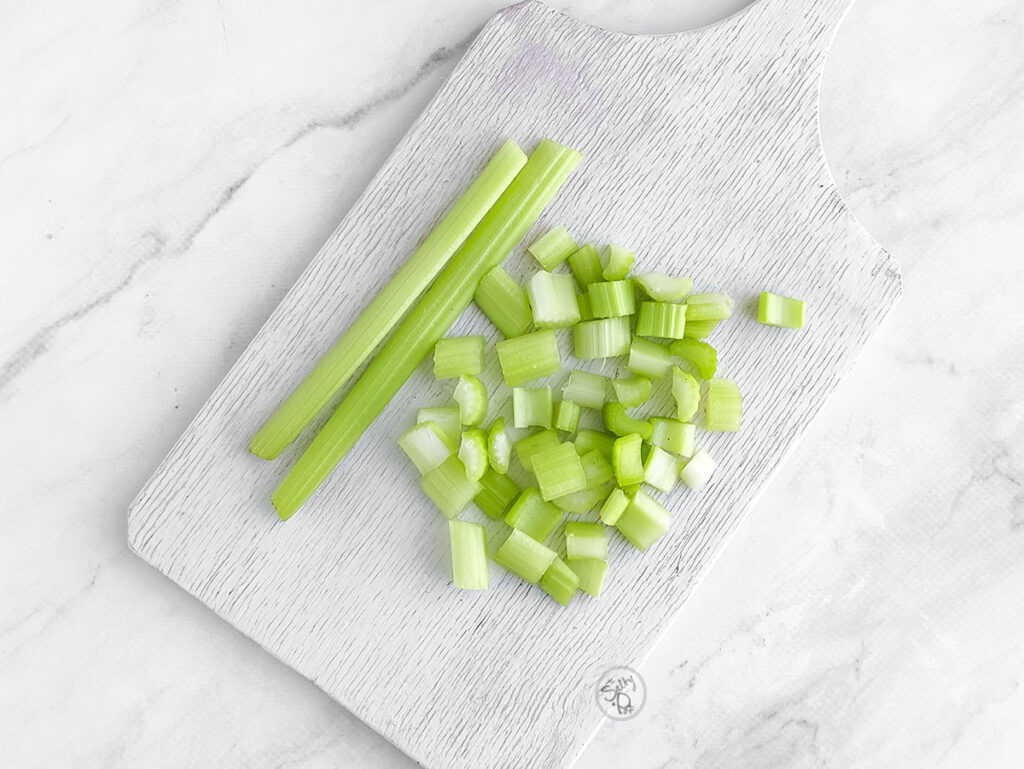 A white wooden cutting board with two whole celery stalks and a pile of freshly diced bright green celery bits.